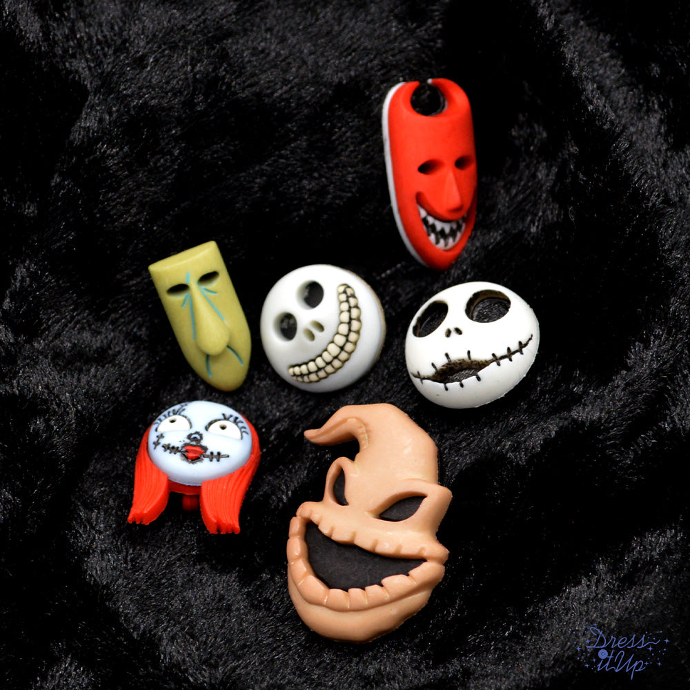 A collection of six Halloween-themed mask buttons are displayed on a black fabric background. The masks have various expressions and colors, including red, green, white, and orange.